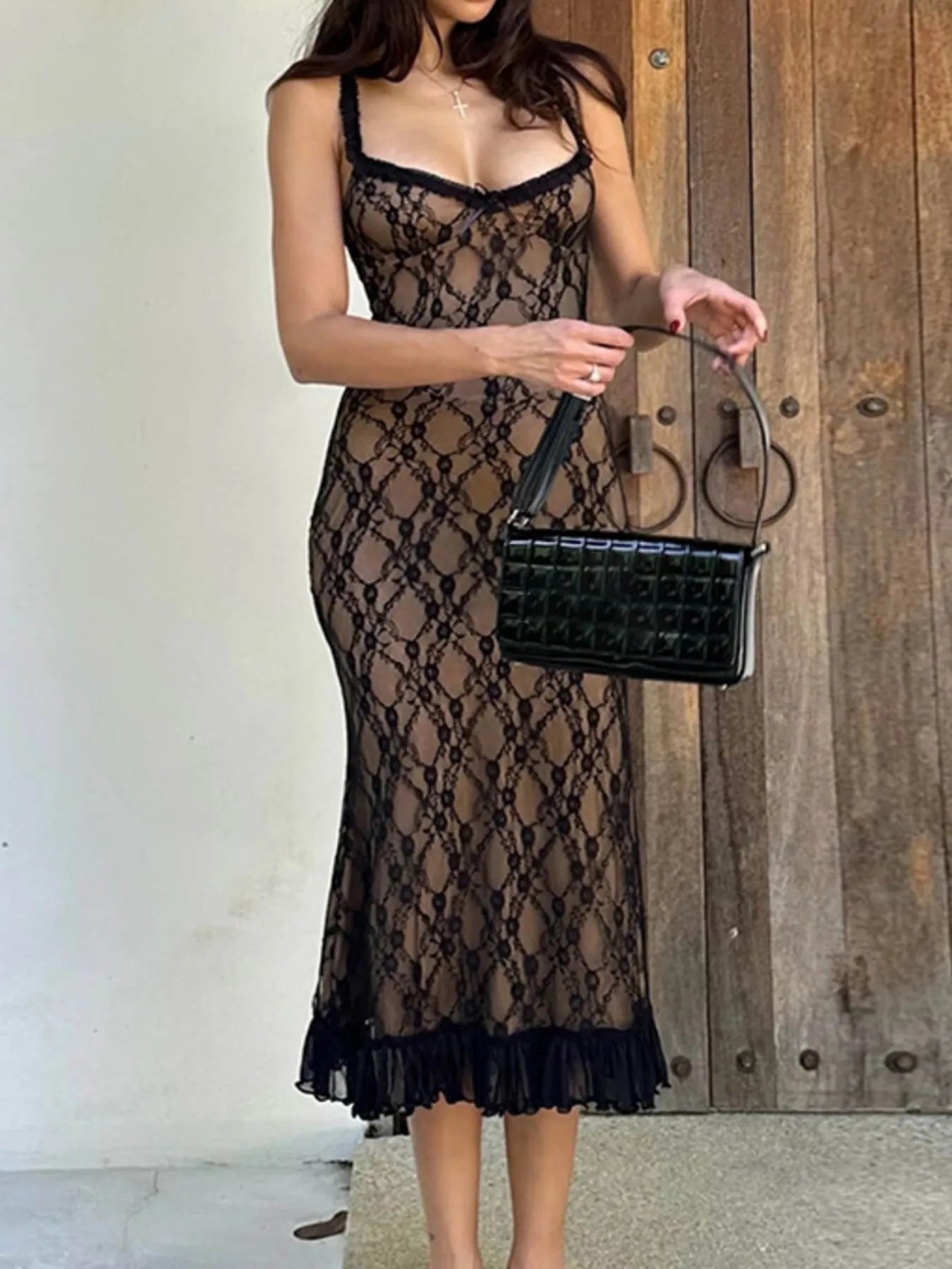 Woman wearing a black lace dress holding a black handbag against a wooden wall.